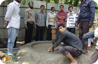 A group of men stands around a concrete tube well being constructed by a mason.
